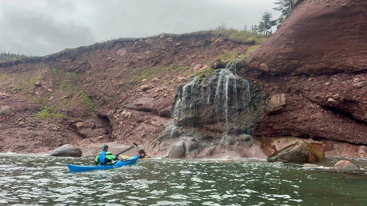 A person in a blue kayak paddling on calm water in front of a rocky shoreline with a small waterfall and reddish-brown cliffs under an overcast sky.