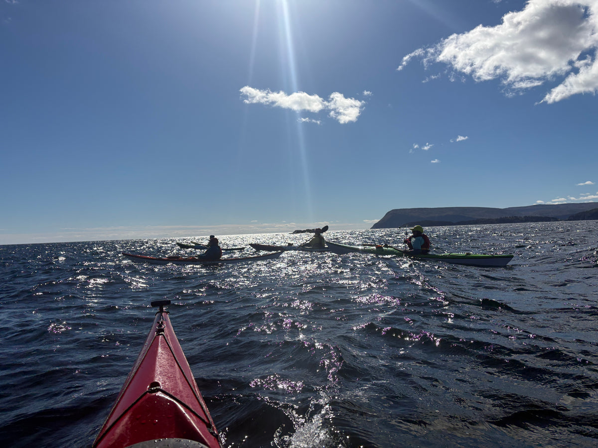A scenic outdoor shot from a kayak on the water under a bright sun with clouds, showing other kayakers on the horizon near Cape Smokey.