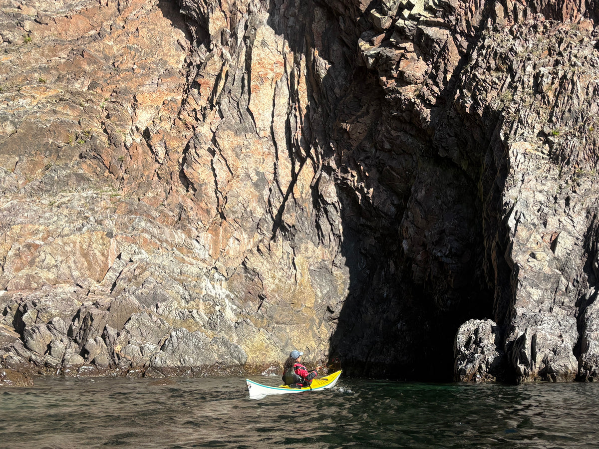 A person kayaking in front of a large rocky cliff and cave formation over dark water.