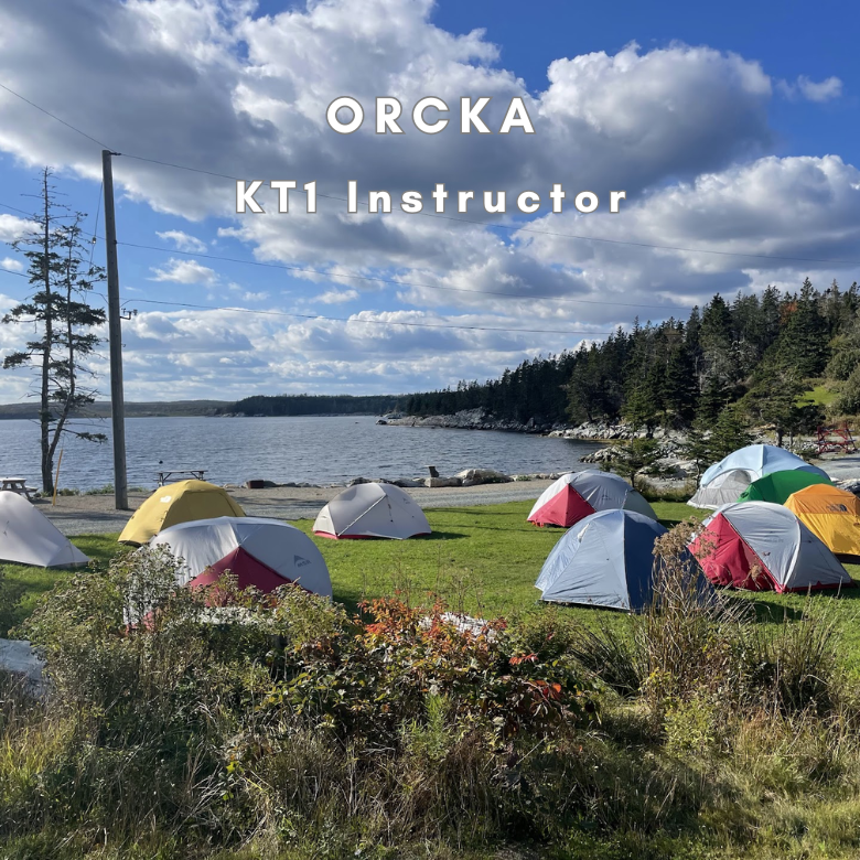 A scenic outdoor campsite with multiple colorful tents pitched on a grassy shore beside a lake under a blue sky with large white clouds. White text overlaid at the top reads 'ORCKA KT1 Instructor'.